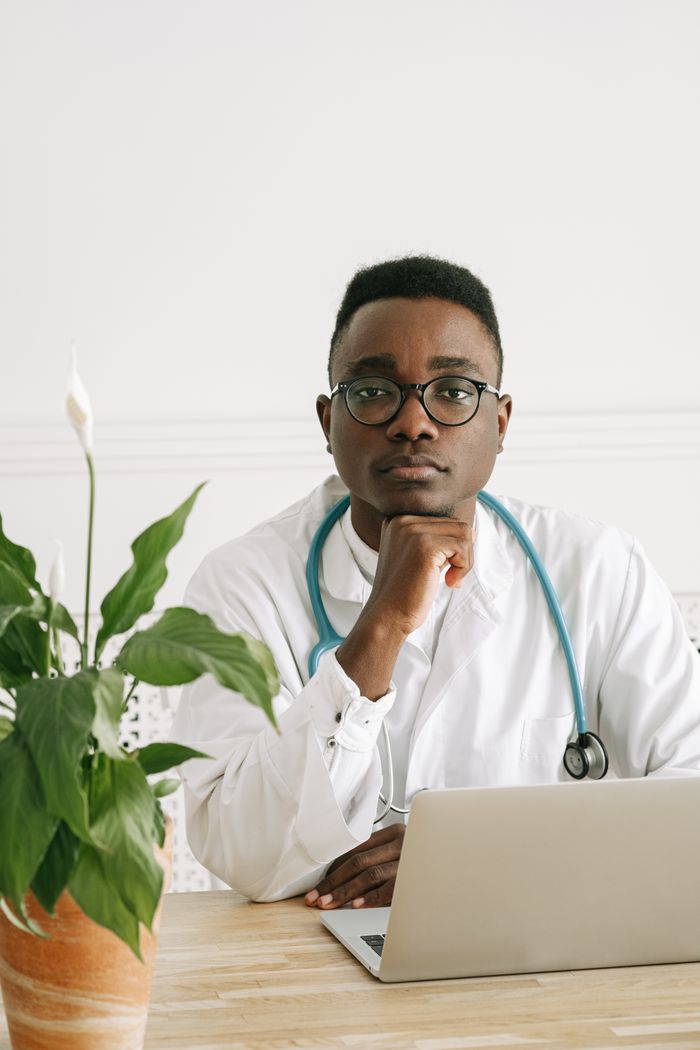 African American doctor sitting at desk with a laptop and plant, wearing a stethoscope, in a calm indoor setting.